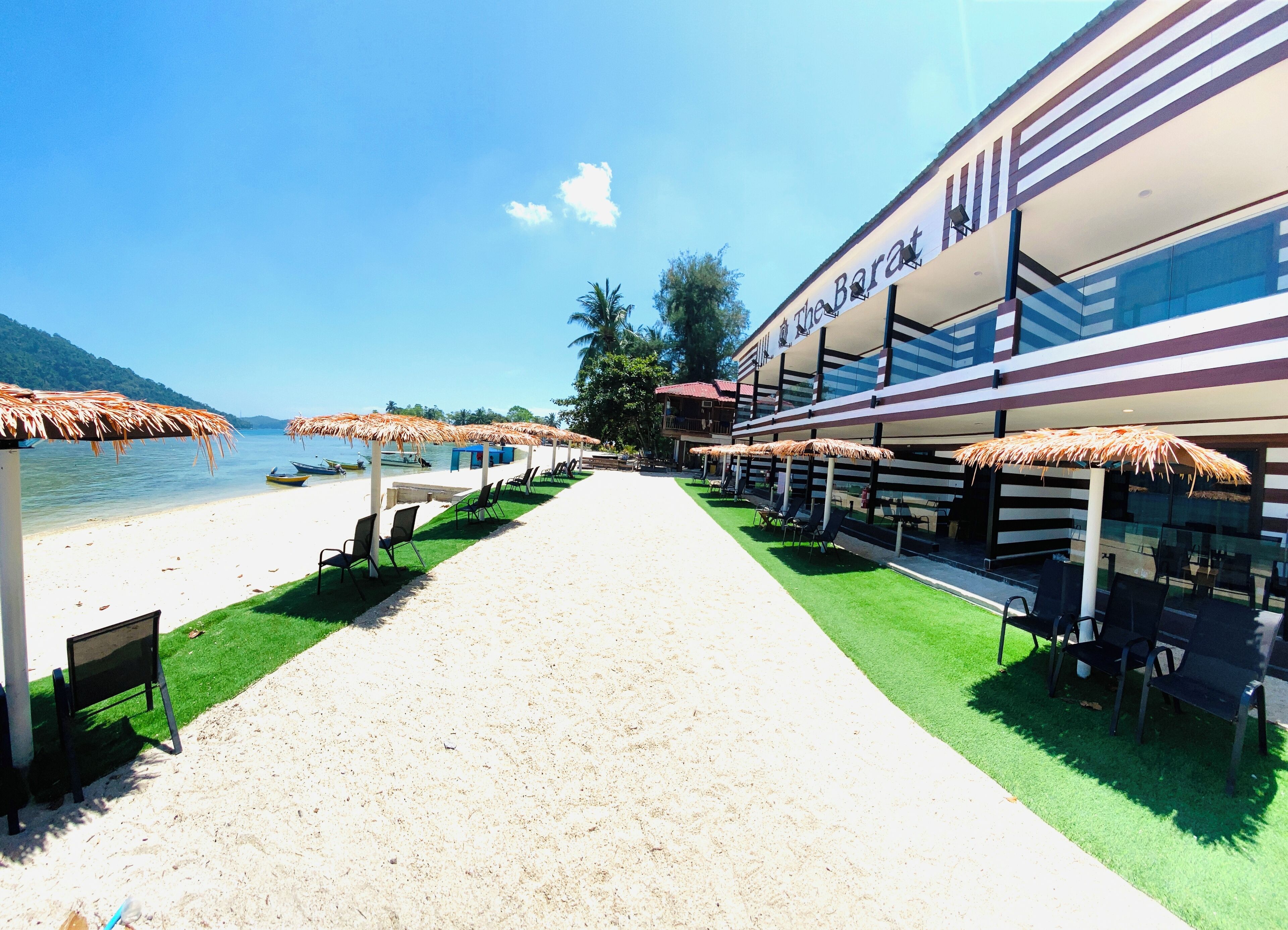 A beachfront hotel with a sandy walkway, thatched umbrellas, and outdoor seating.