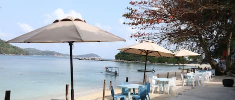 A beachside café with outdoor seating, tables, and umbrellas, overlooking a calm sea and distant hills.