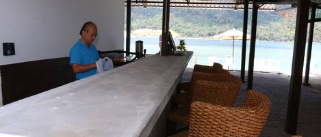 A bar area with a view of the sea, wicker chairs, and a person standing by the counter.
