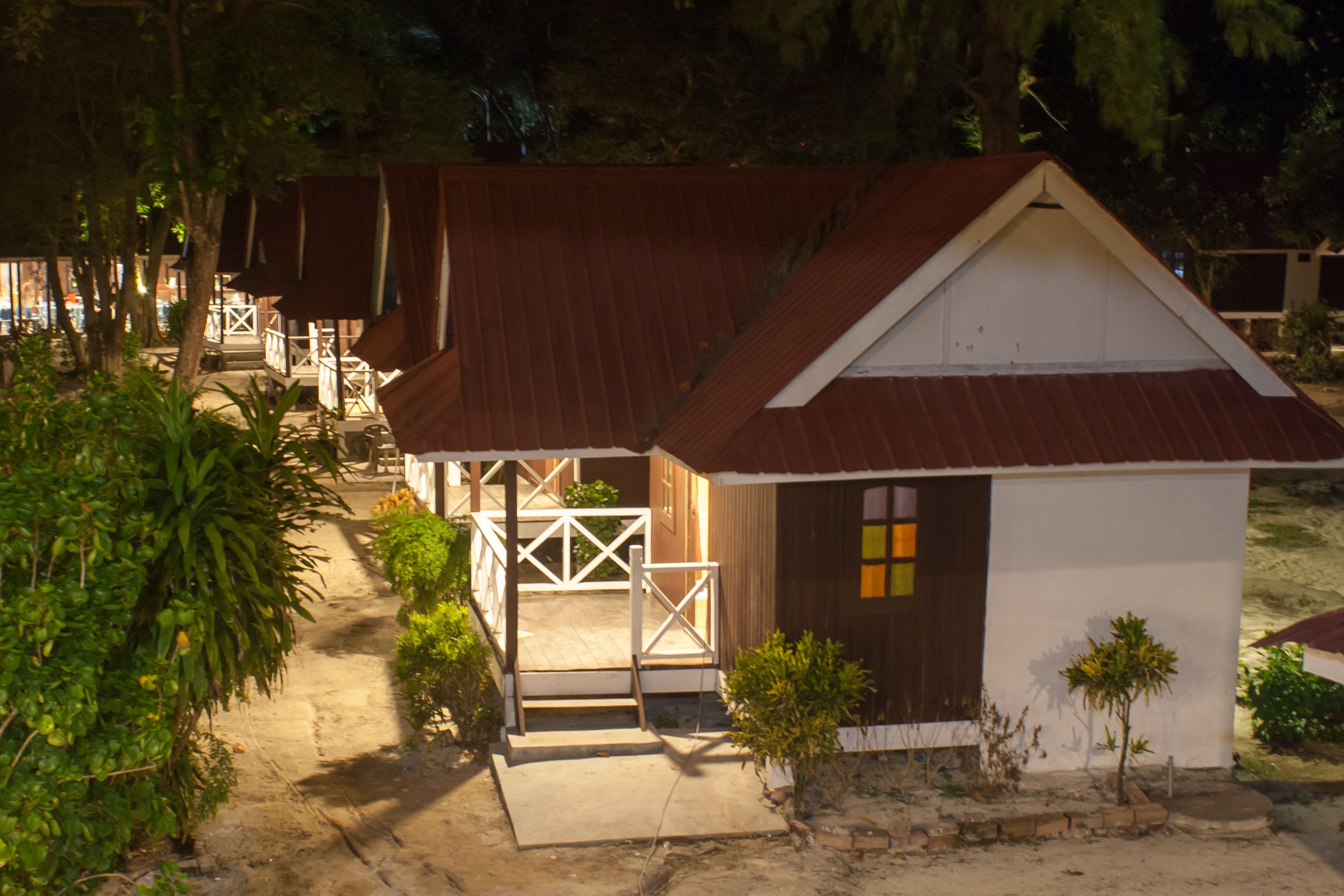 A small, single-story building with a white facade and a brown roof, surrounded by greenery and a wooden deck.