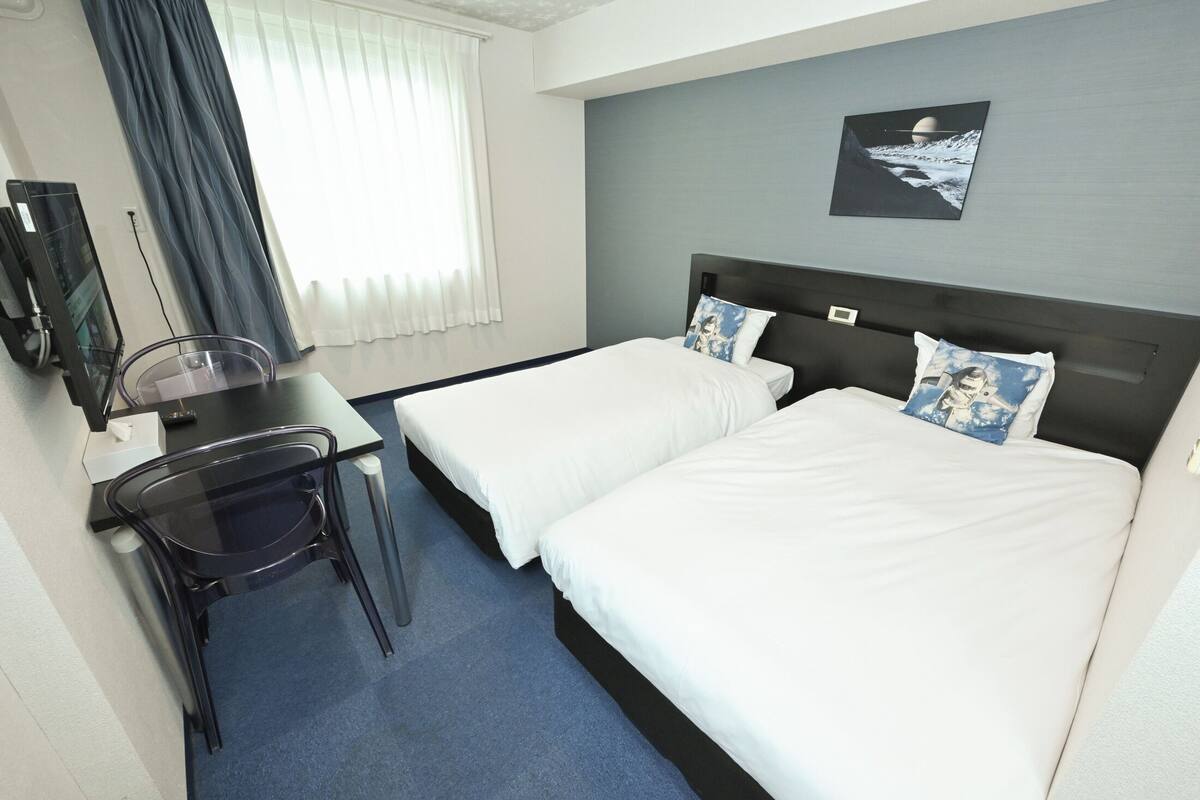A hotel room with two beds, a desk with a chair, a TV, and a window with curtains.