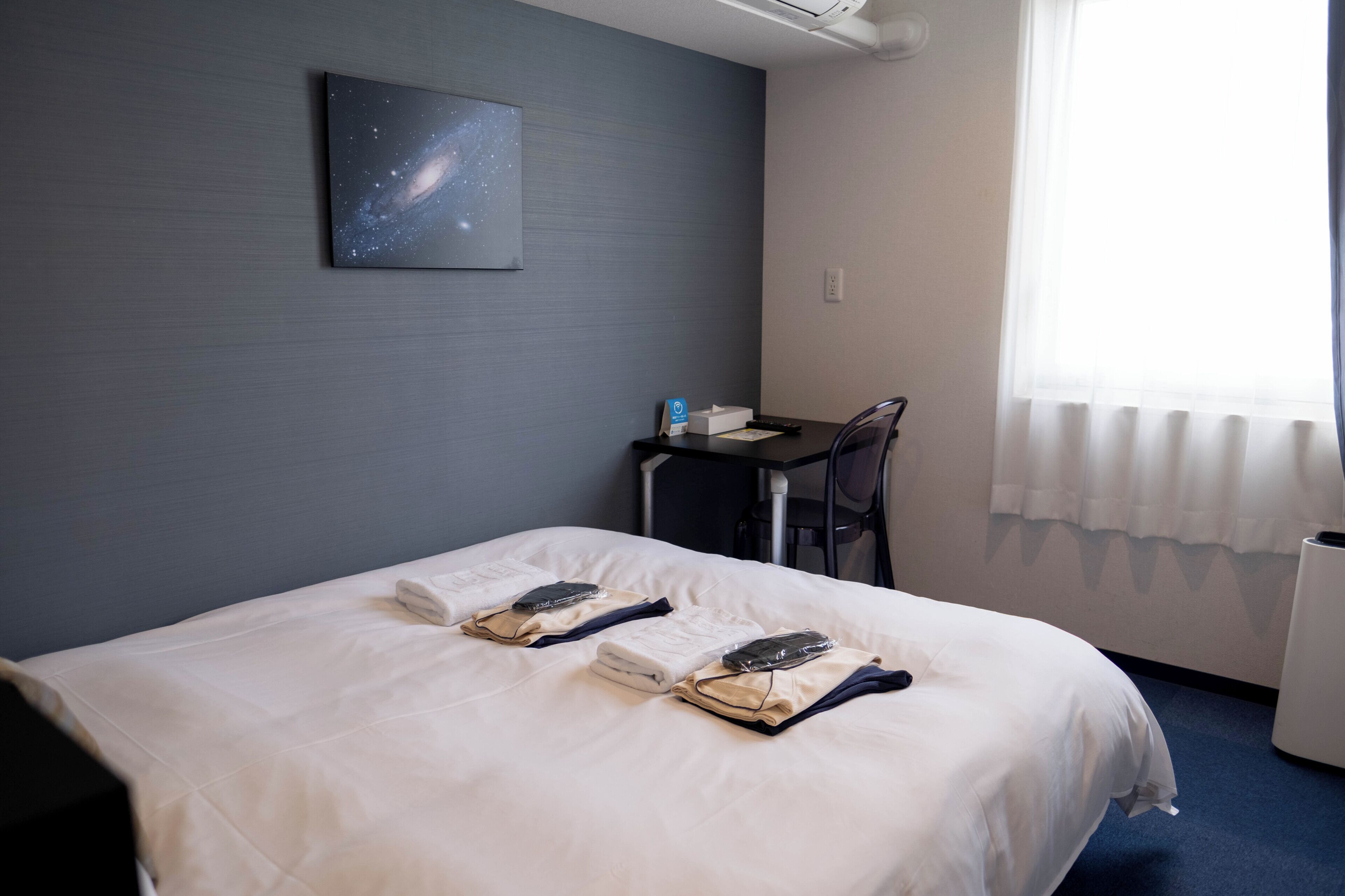 Una habitación de hotel con una cama, un escritorio, una silla, un ventanal con cortinas y un cuadro con temática de galaxia en la pared.