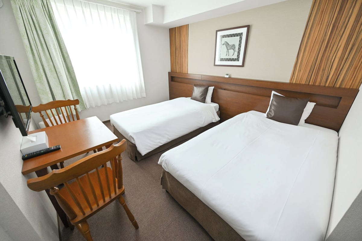A hotel room with two single beds, a wooden desk with a chair, a television, and a framed picture on the wall.
