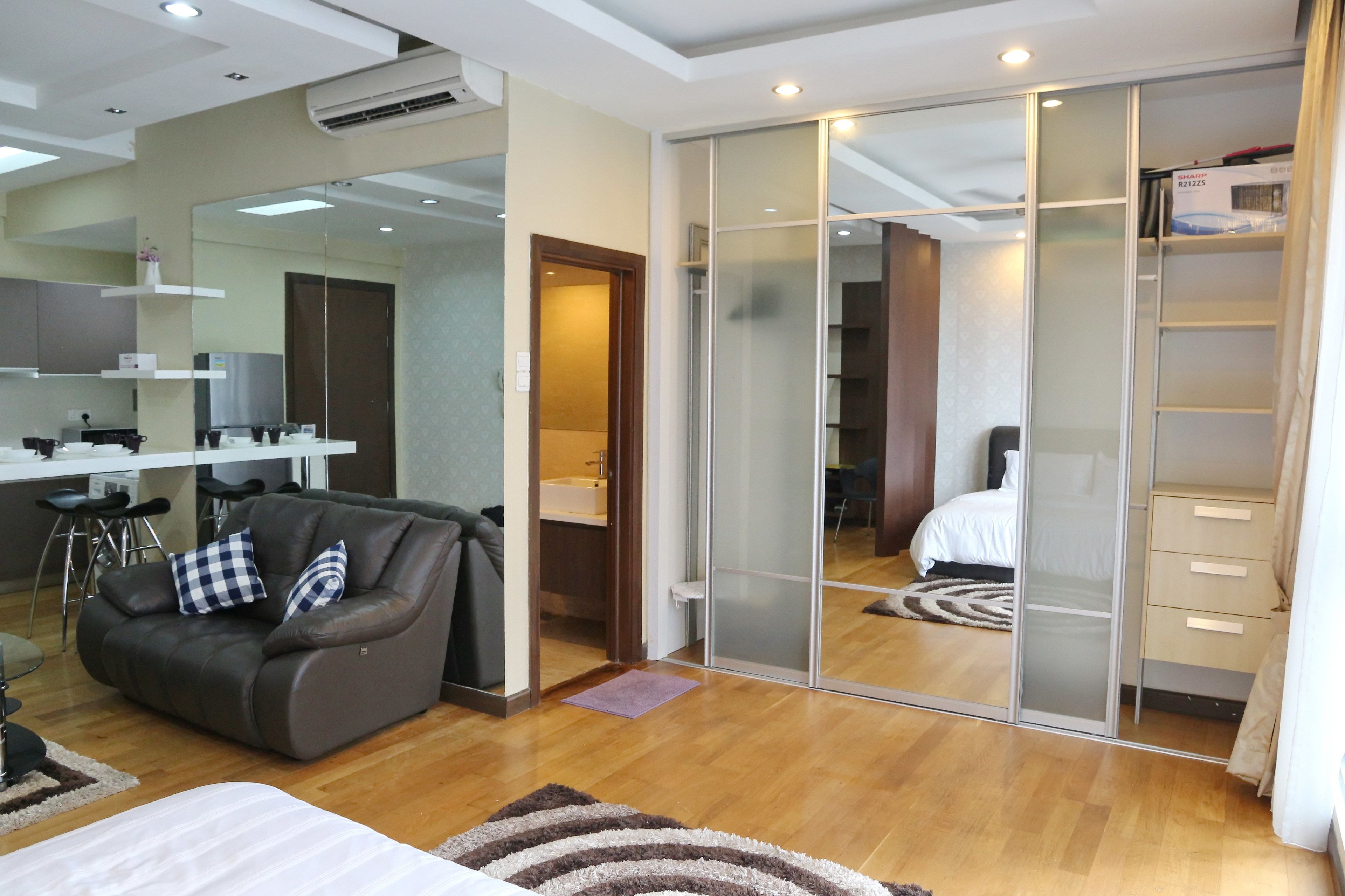 A modern hotel room with a sliding glass door closet, a sofa with cushions, a small kitchen area, and a bed.
