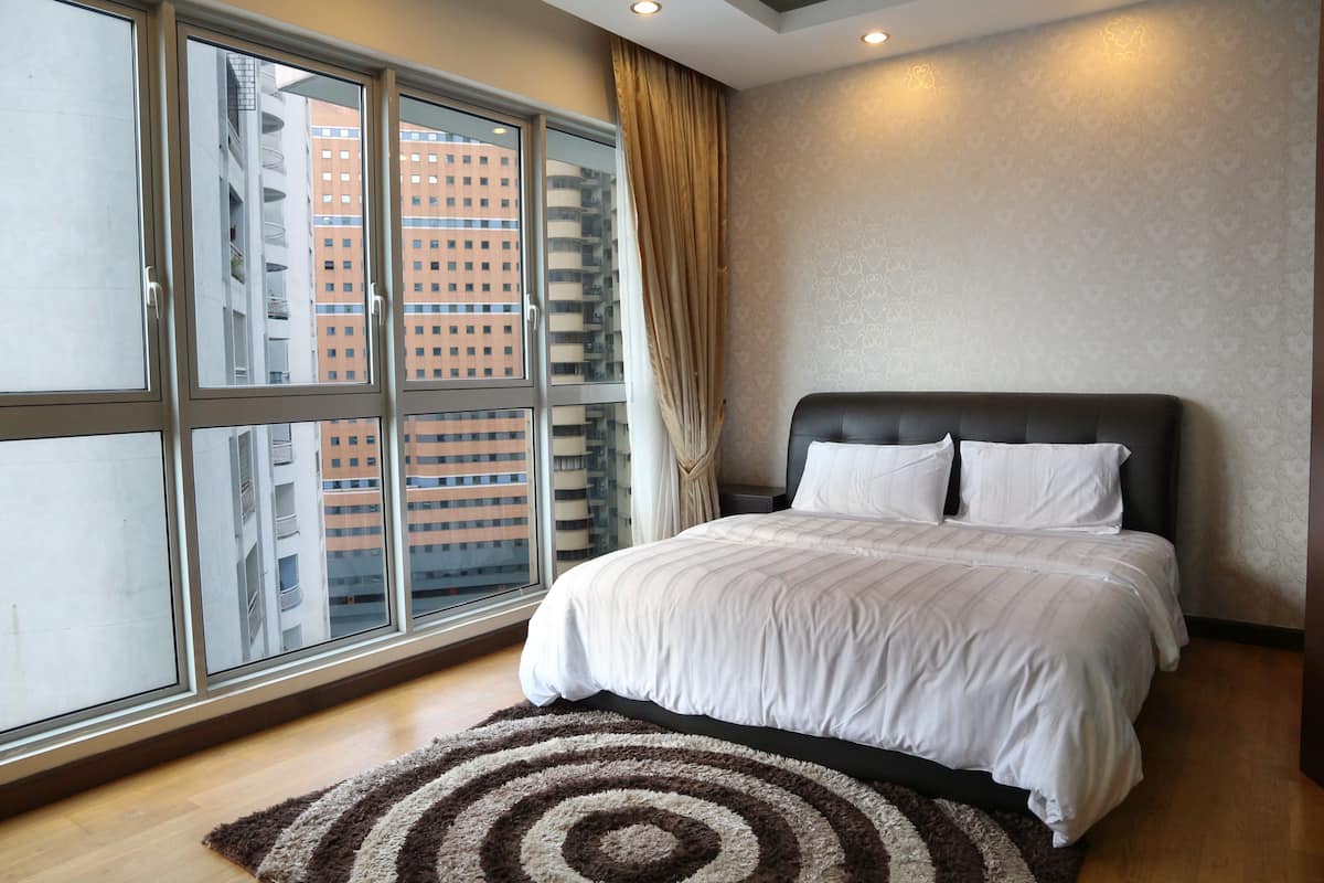 A bedroom with a large window, a bed with white bedding, a brown rug, and a curtain.