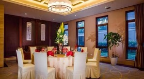 Un coin repas avec une table ronde dressée pour six personnes, entourée de chaises, avec une grande fenêtre et une plante en pot en arrière-plan.