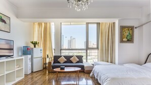 Une chambre d’hôtel moderne avec un plancher en bois, un canapé, un lit, une télévision, un distributeur d’eau et une grande fenêtre donnant sur la ville.