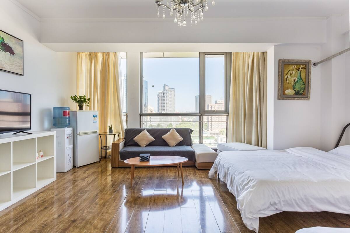 Una habitación de hotel moderna con piso de madera, un sofá, una cama, un televisor, dispensador de agua y un ventanal con vistas a la ciudad.