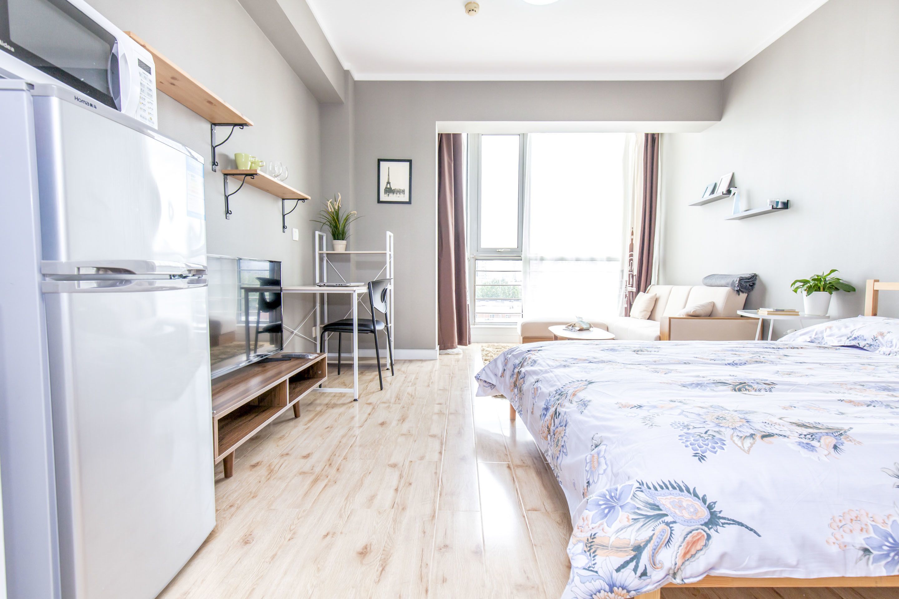 A modern hotel room with a bed, a desk, a chair, a refrigerator, and a window with curtains.