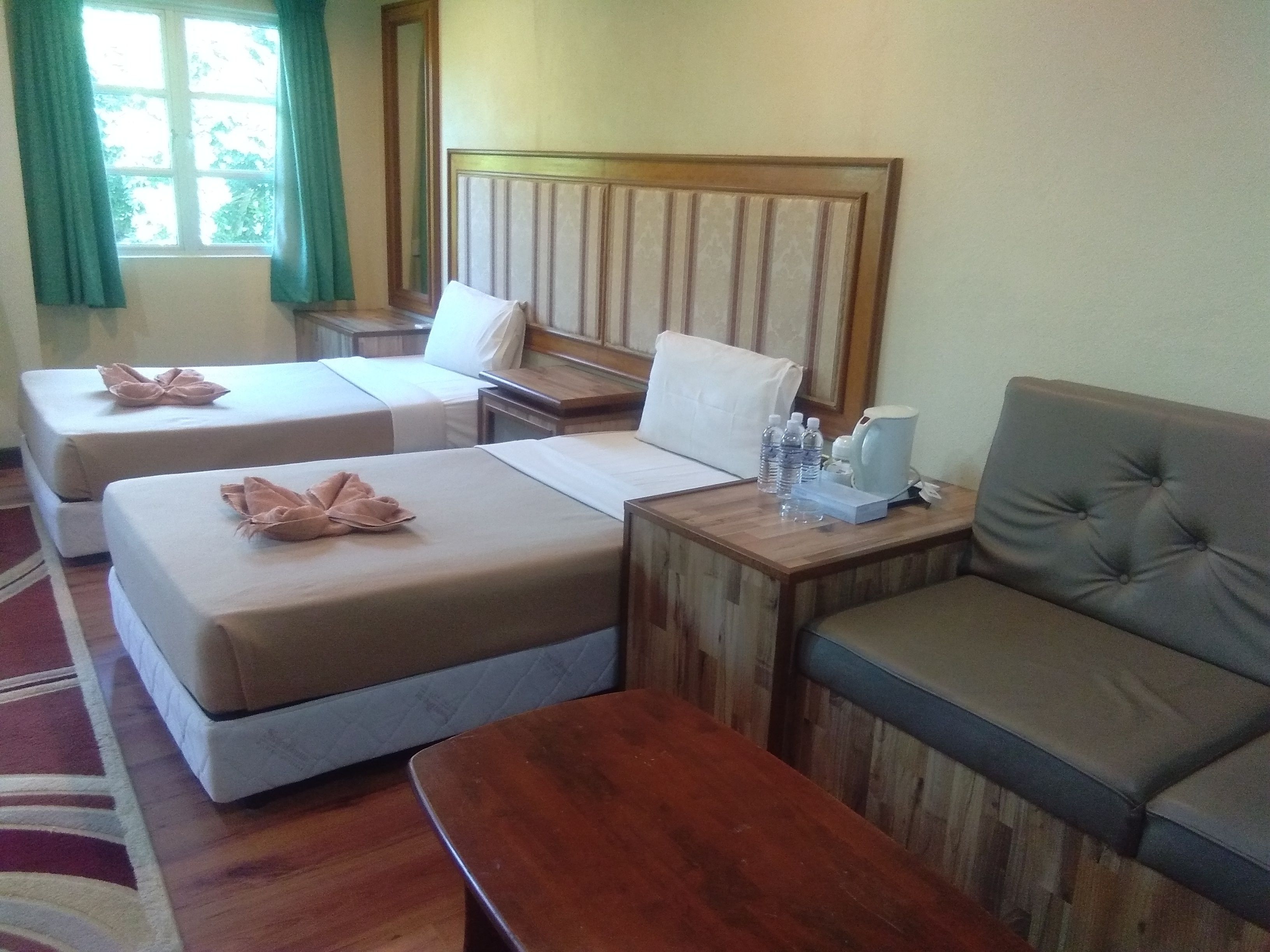 A hotel room with two single beds, a sofa, a wooden nightstand, and a window with curtains.