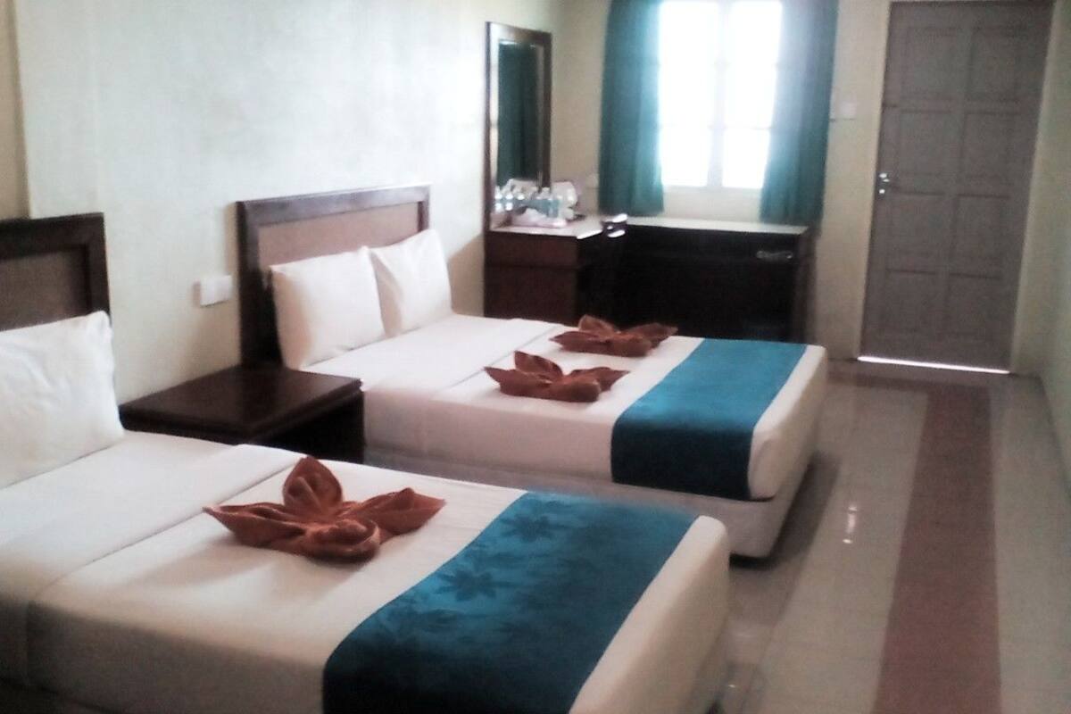 Habitación de hotel con dos camas, cada una con colcha azul y blanca, y cabeceros de madera. Se observa un mesita de noche con una lámpara y un espejo en la pared.