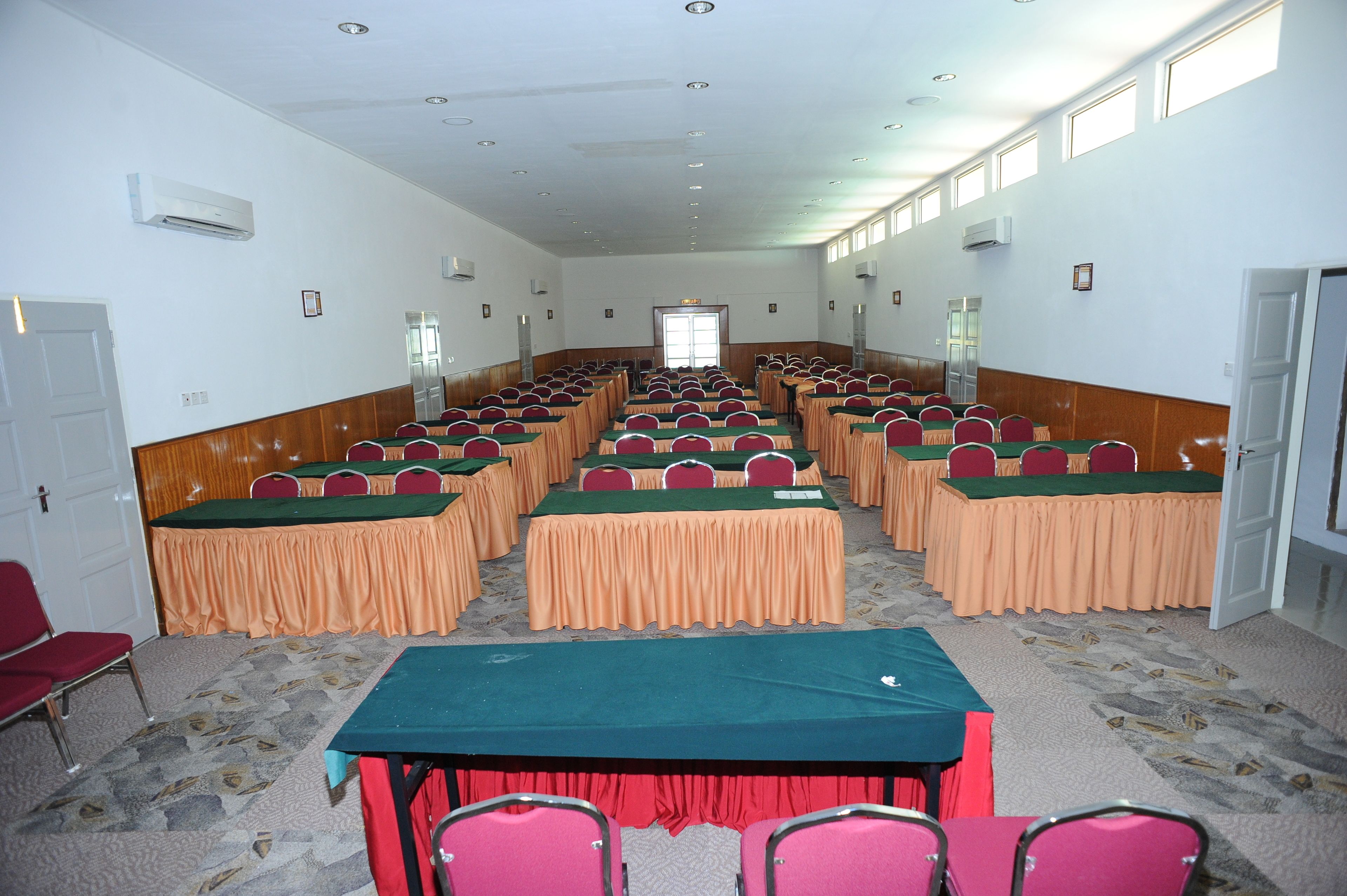 A conference room with rows of tables and chairs, air conditioning units on the walls, and a door at the far end.