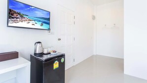 A hotel room with a large TV displaying a beach scene, a small refrigerator with a kettle and glasses, and a white door. - The Way Phuket Resort (Thep Krasattri)