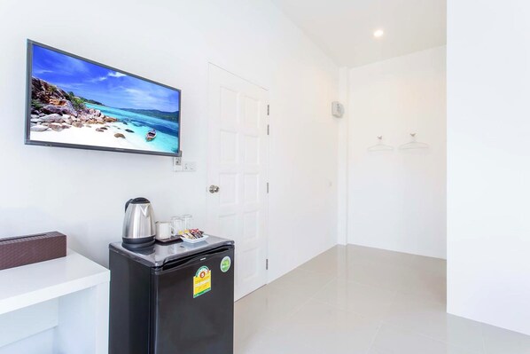 A hotel room with a large TV displaying a beach scene, a small refrigerator with a kettle and glasses, and a white door. - The Way Phuket Resort (Thep Krasattri)