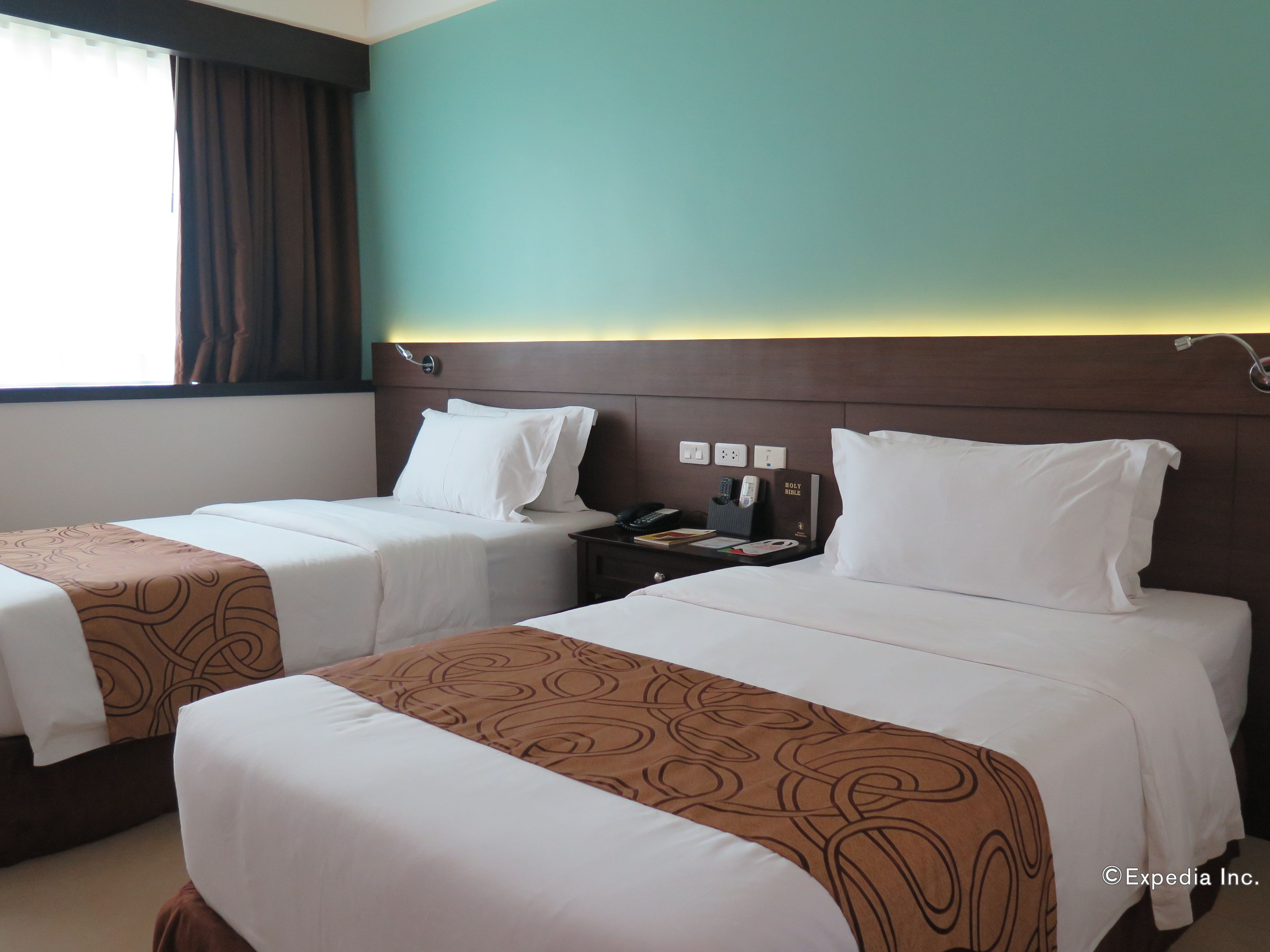 A hotel room with two beds, a nightstand with a phone and a Bible, and a window with curtains.