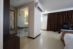 A modern hotel room with a bed, a TV, a desk, and a bathroom with a glass shower enclosure. - Kew Hotel (Tagbilaran)