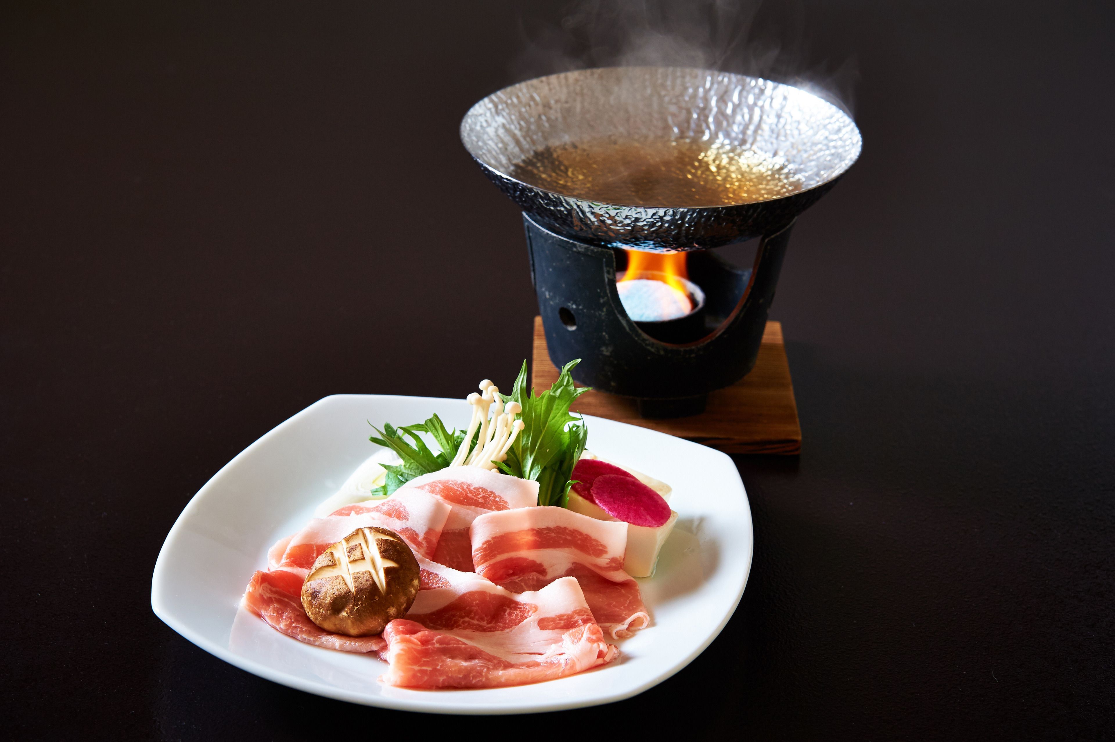 Un plato de comida con diferentes carnes y verduras, una olla caliente con vapor saliendo, y una mesa de madera.
