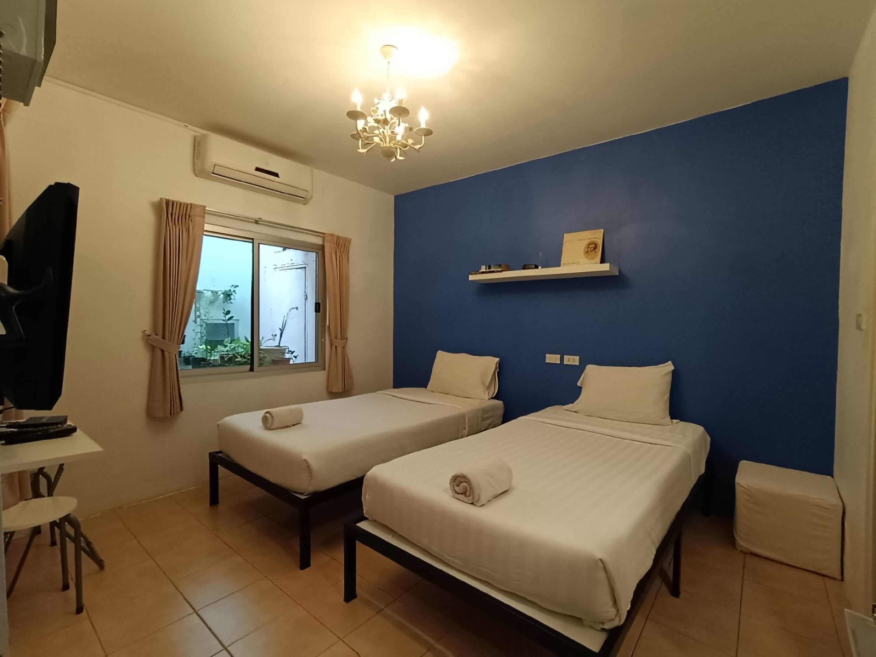 A room with two single beds, a desk with a chair, a small table, and a window with curtains.