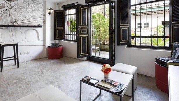 A modern living room with a marble floor, a white sofa, a coffee table with a vase, and a red ottoman. There are open black-framed windows with a view of greenery outside. - Chino Town Gallery Alley (Phuket)
