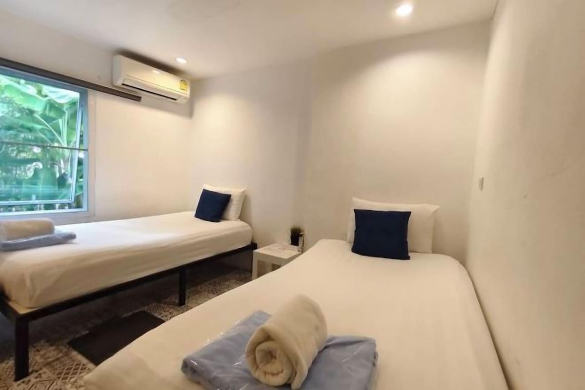 Habitación de hotel con dos camas, una mesita de noche pequeña, un equipo de aire acondicionado y una ventana con vistas.