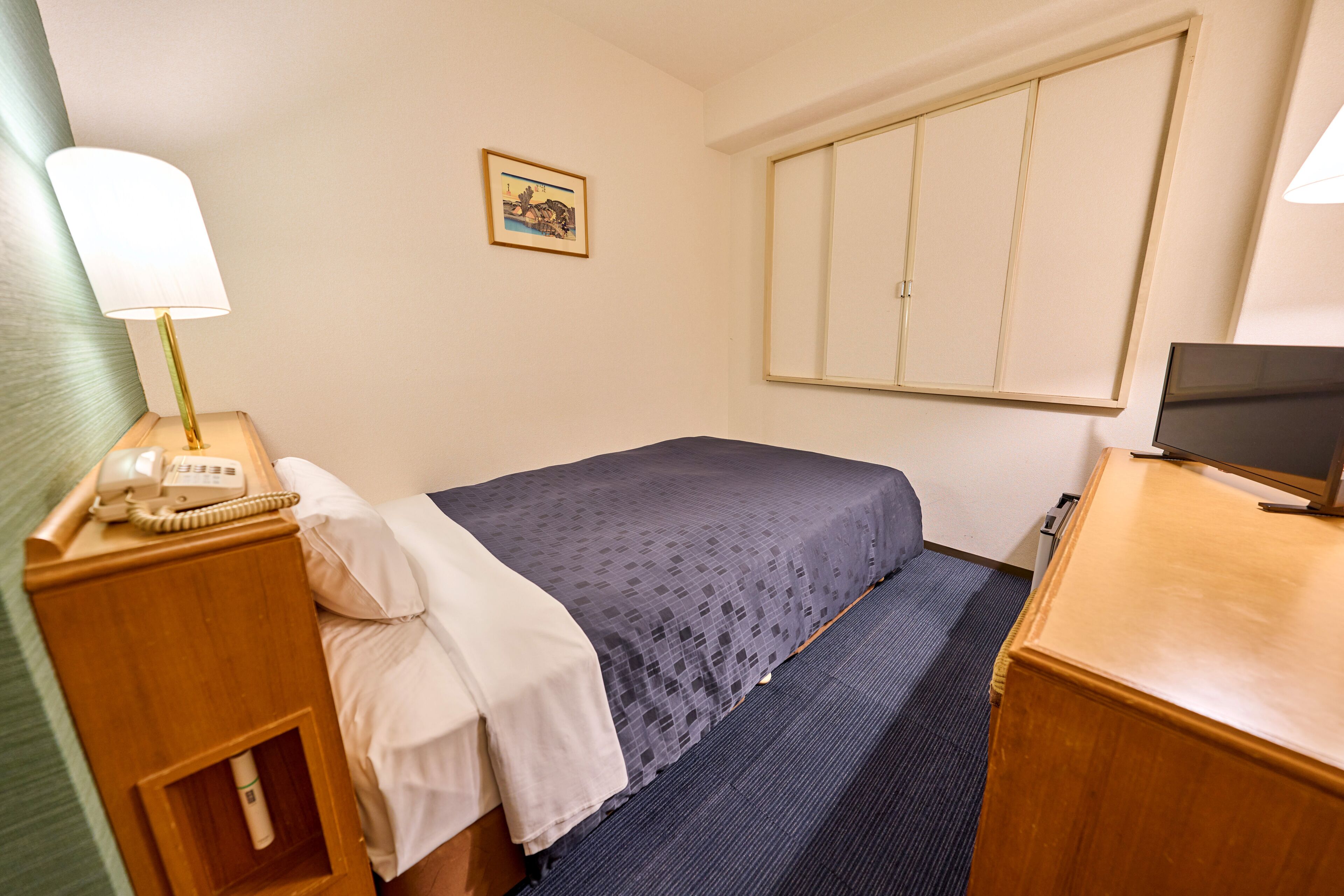 Habitación de hotel con cama, escritorio con lámpara, televisor, teléfono y un cuadro en la pared.