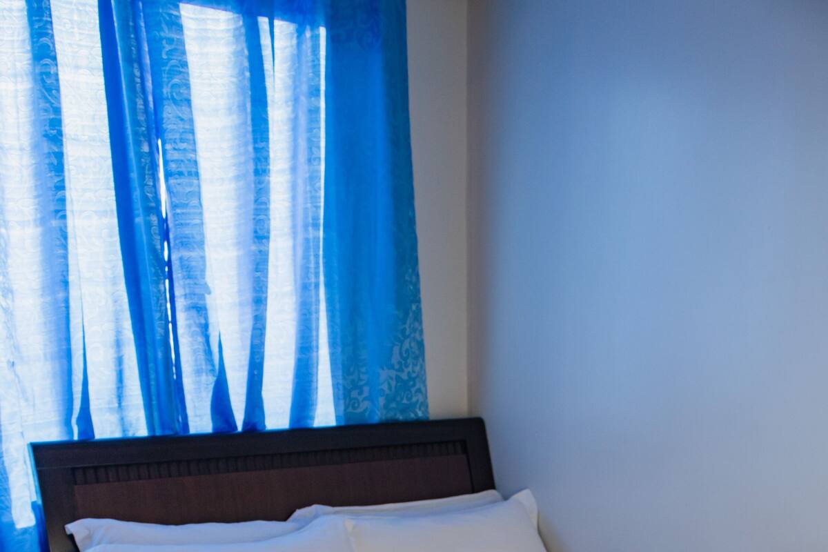 A neatly made bed with white linens and pillows, a wooden headboard, and a window with blue curtains.