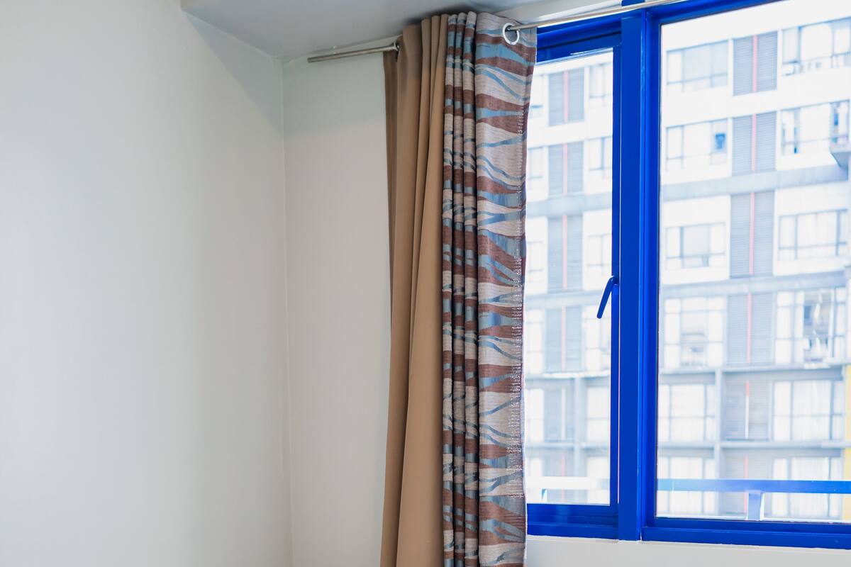 A single bed with a white mattress and a rolled white towel, a window with blue trim, and a curtain with a striped pattern.