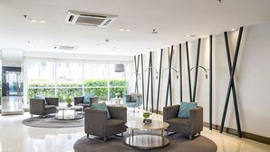 En moderne lobby med grå sofaer, runde borde og et stort vindue med udsigt over grøn vegetation.