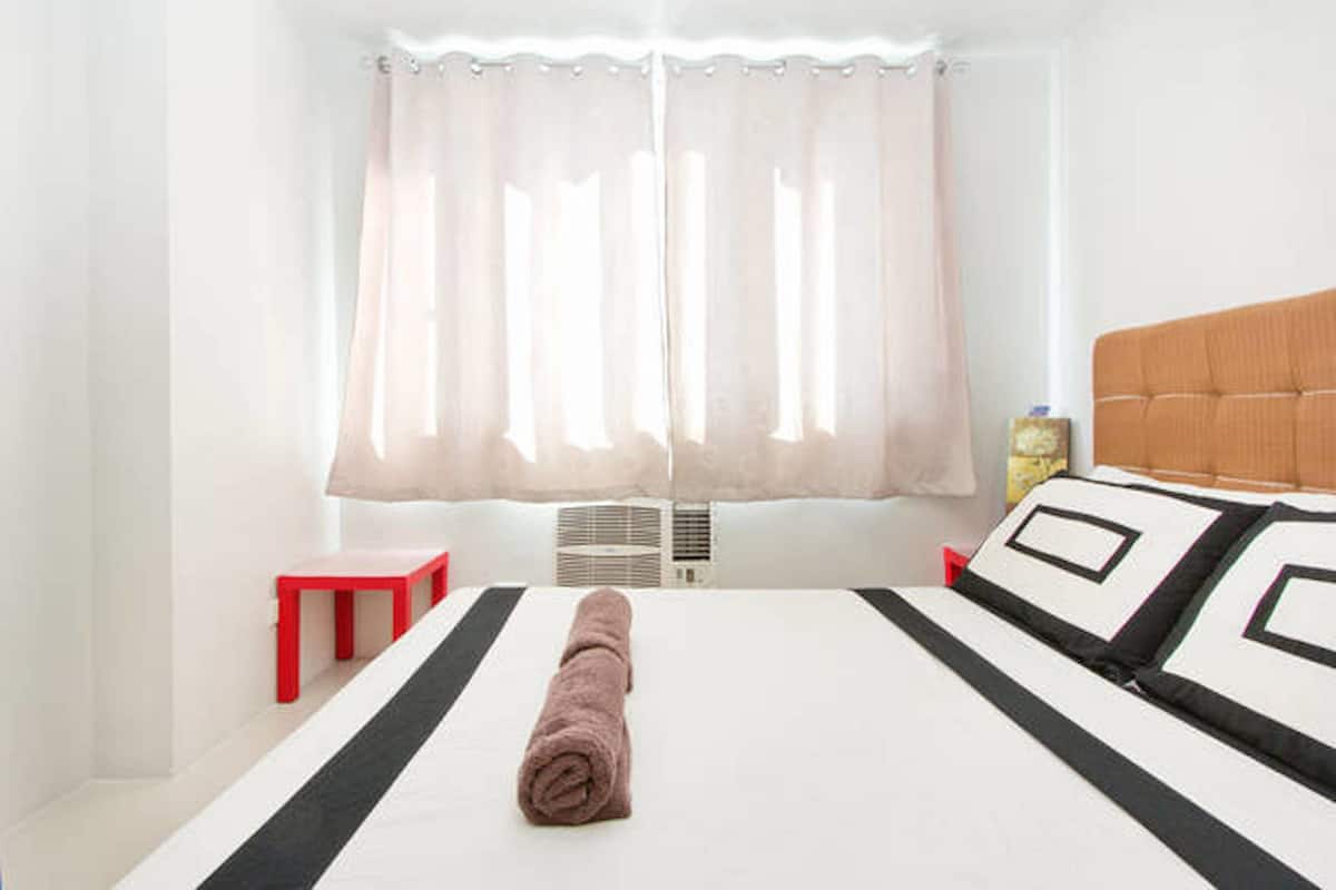 A hotel room with a bed, a red stool, a window with sheer curtains, and a bottle on the nightstand.