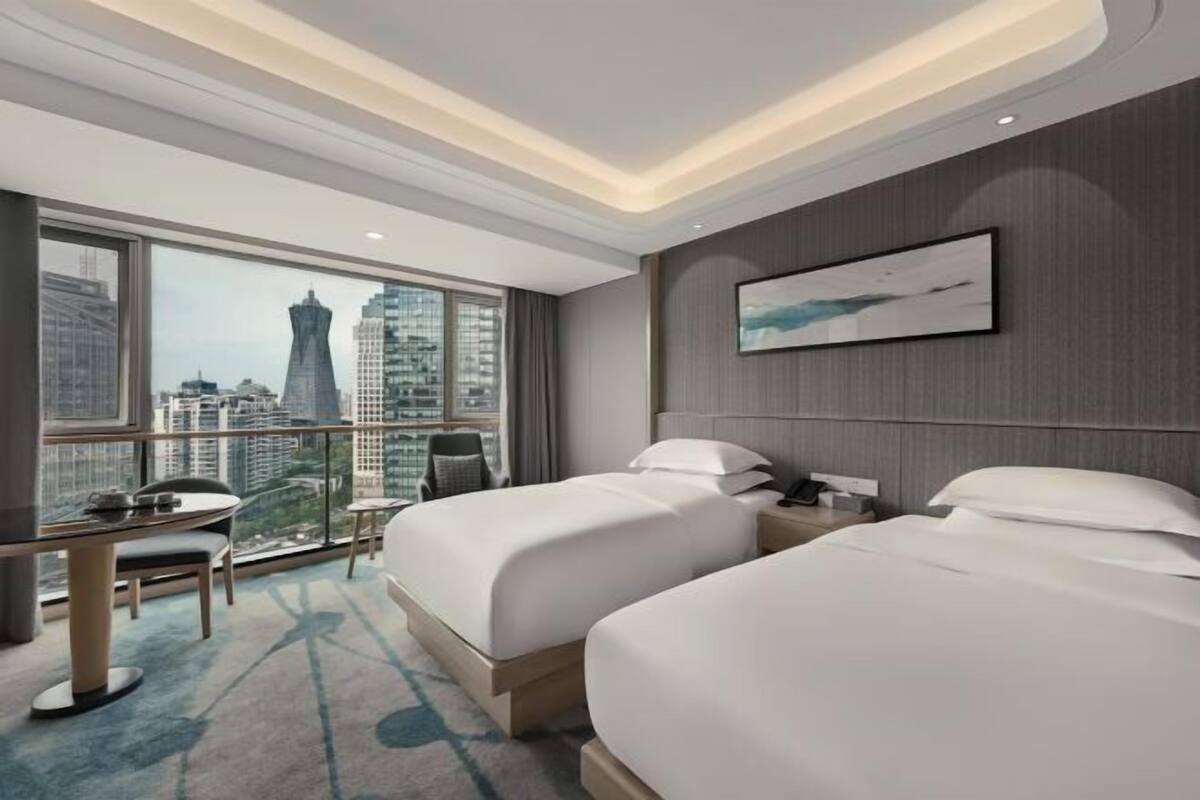 A hotel room with two beds, a desk, a chair, and a large window with a city view.