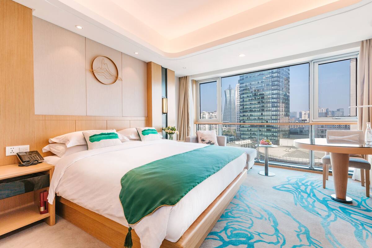 A modern hotel room with a large bed, a desk, a chair, and a view of the cityscape through large windows.