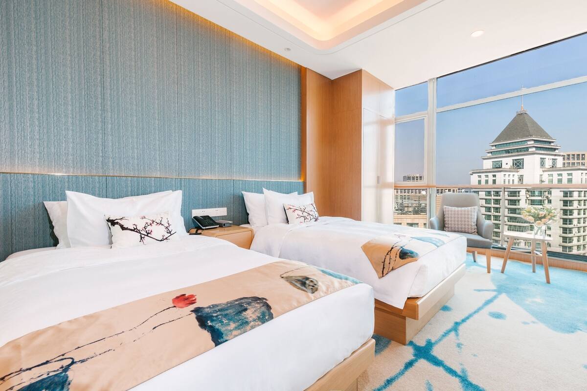 A hotel room with two beds, a large window with a city view, and a balcony with a chair.