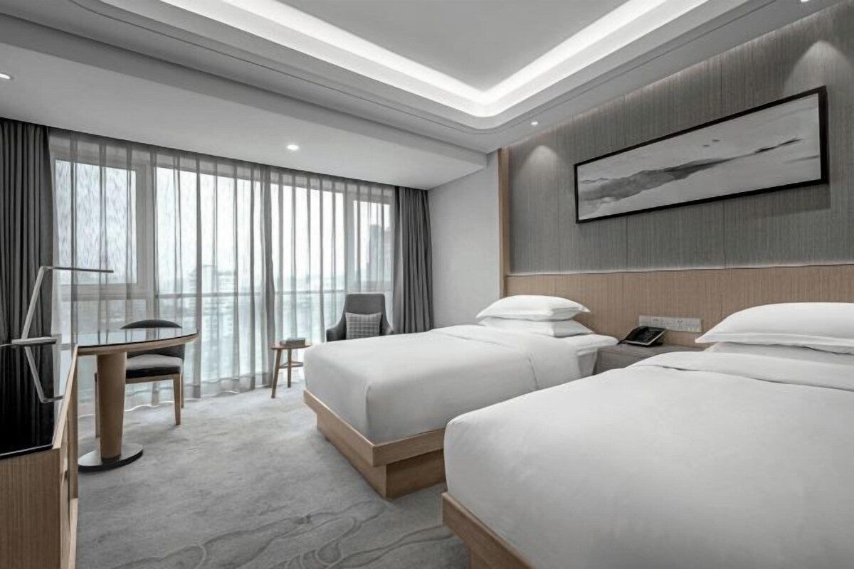 A modern hotel room with two beds, a desk, a chair, and a large window with curtains.