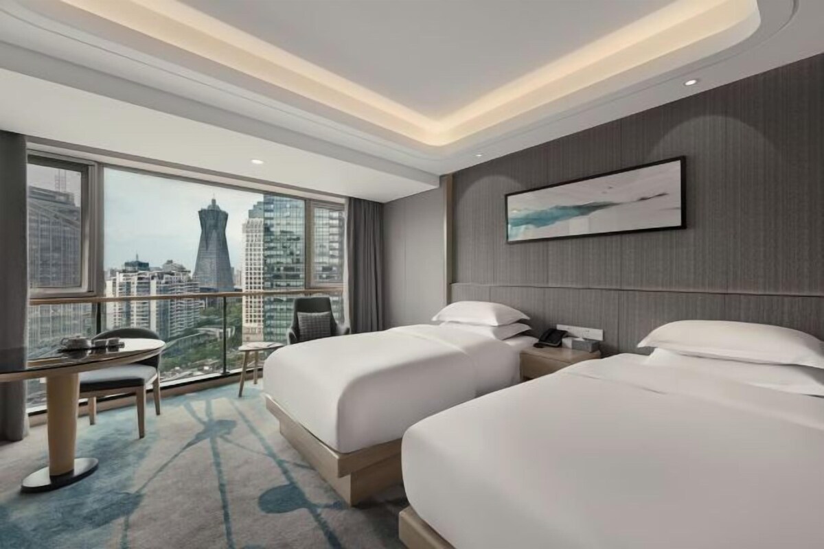 A hotel room with two beds, a desk, a chair, and a large window with a city view.