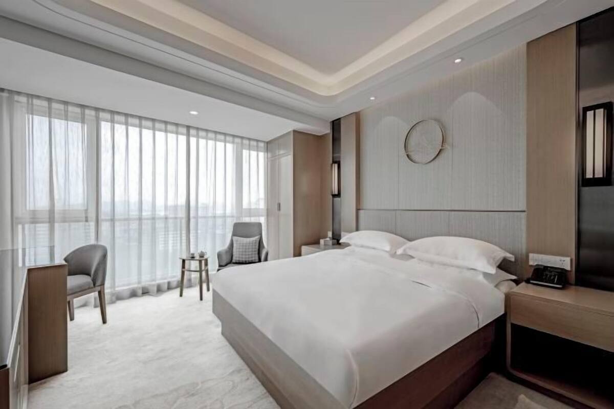 A modern hotel room with a large bed, two bedside tables, a chair, and a large window with sheer curtains.