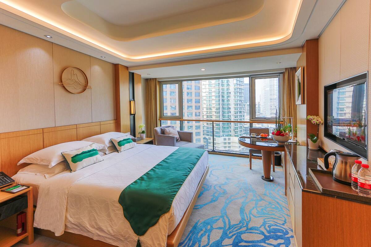 A hotel room with a large bed, a desk with a TV, a balcony with a view of tall buildings, and a modern design.