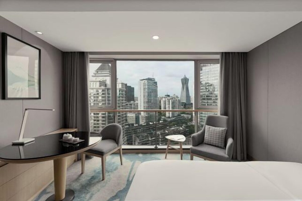 A modern hotel room with a city view, a desk, two chairs, a small table, and a bed.