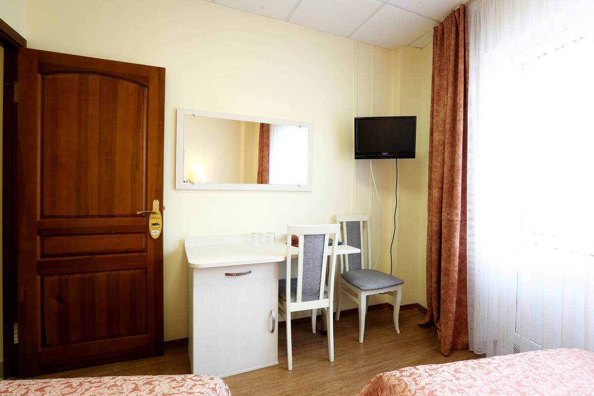 A hotel room with a wooden door, a desk with a mirror, a television, two chairs, and a bed.