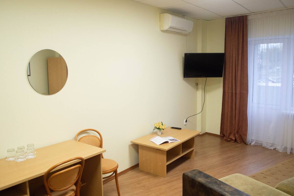 A room with a round mirror, a TV mounted on the wall, a wooden desk with a chair, a small table with a vase of flowers, and a sofa.
