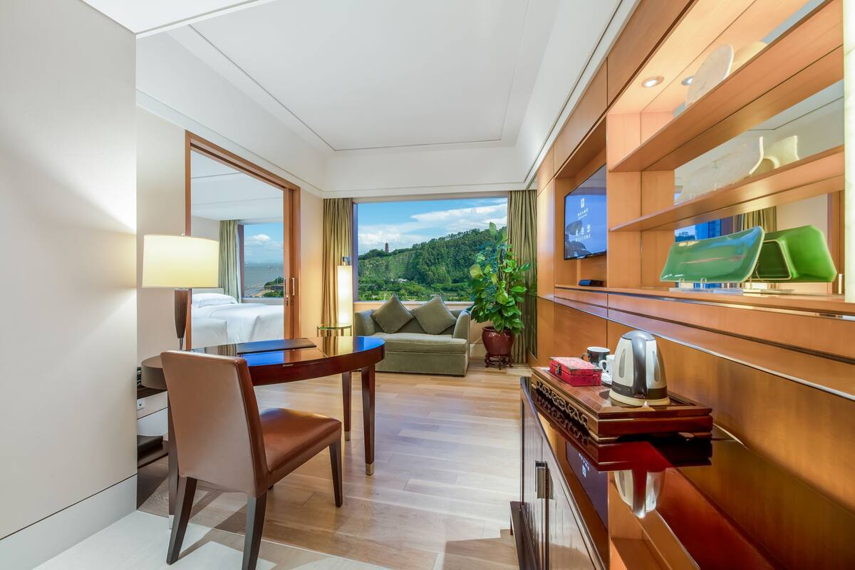 Ein Hotelzimmer mit Bett, Schreibtisch, Stuhl, Sofa, Fernseher und Blick auf eine grüne Landschaft.
