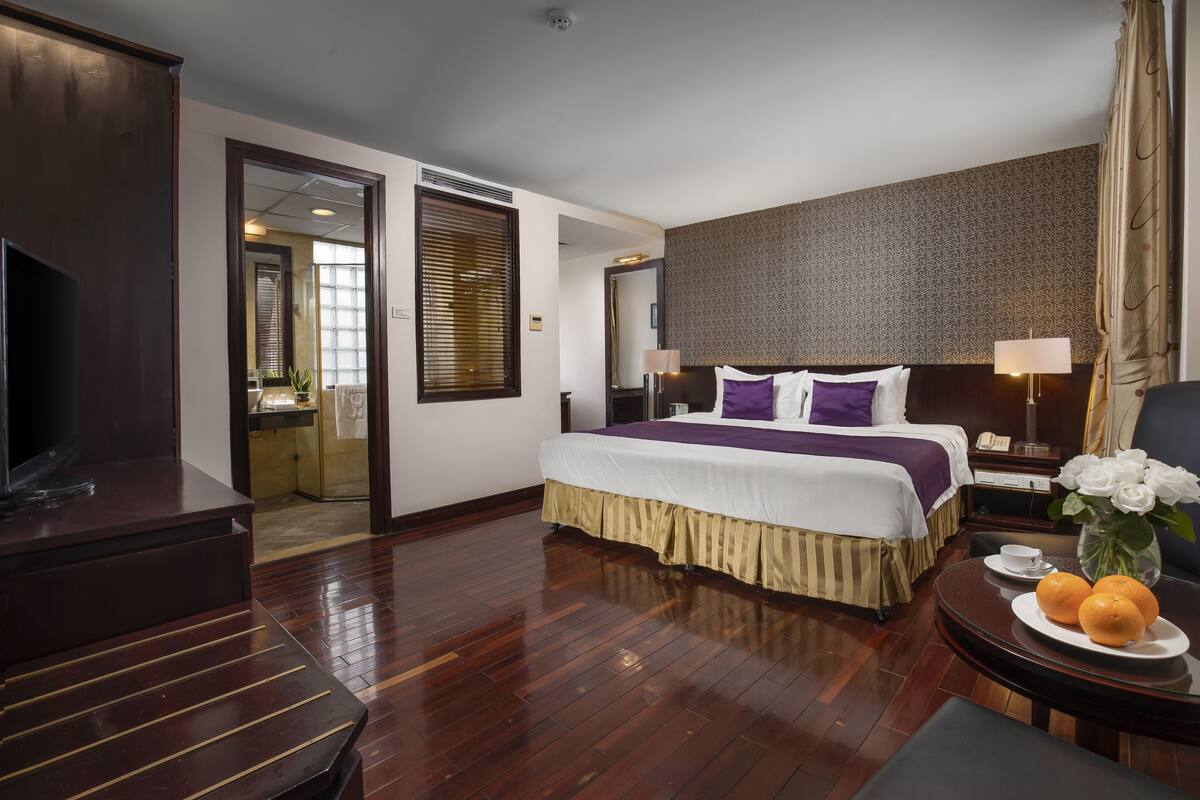 A hotel room with a large bed, a television, a desk with a lamp, and a vase with flowers.