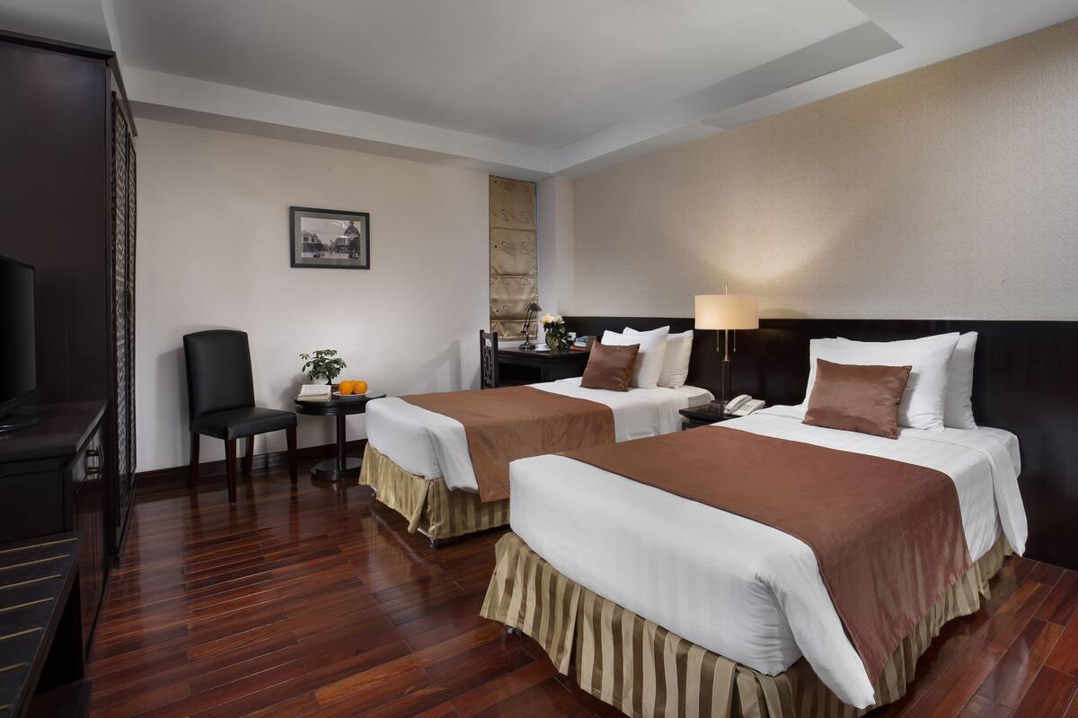 A hotel room with two beds, a desk, a chair, a TV, and a lamp.
