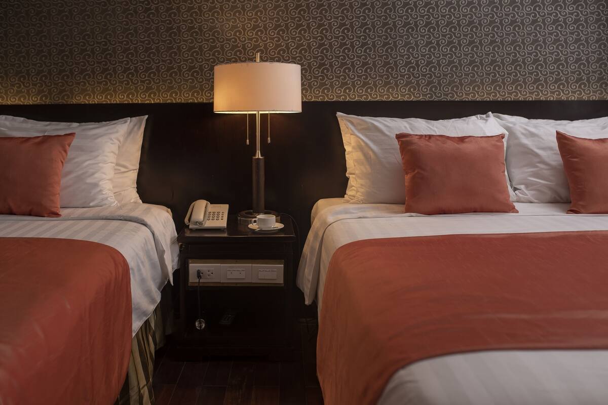 A hotel room with two beds, a nightstand with a lamp, a phone, and a cup.