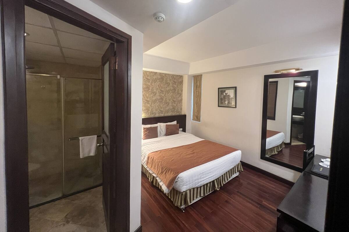 A hotel room with a bed, a mirror, a shower, and a door leading to a bathroom.
