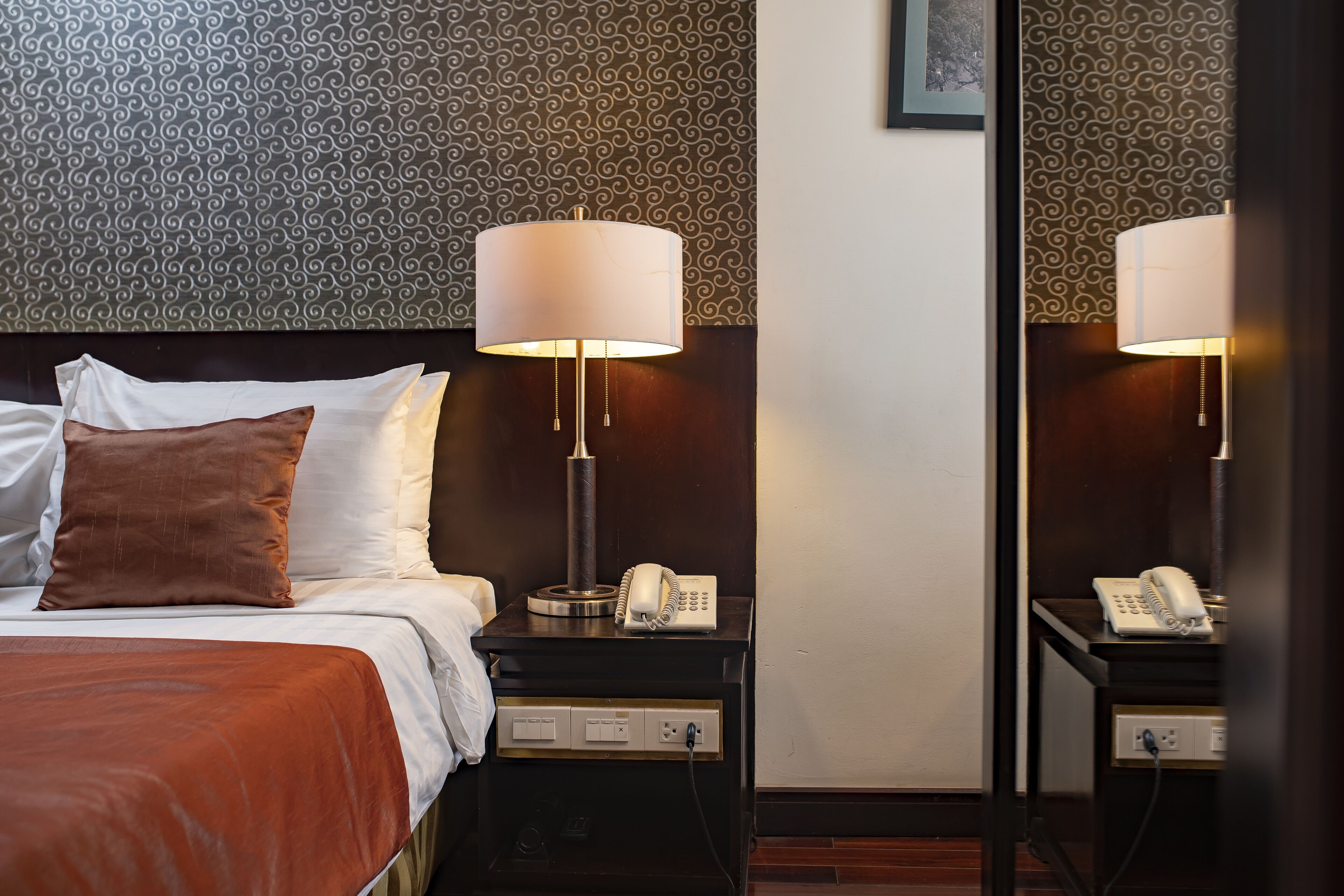 A neatly made bed with a brown pillow, a white pillow, and a white blanket. There is a bedside table with a lamp, a telephone, and a power outlet. The wall has a patterned wallpaper.