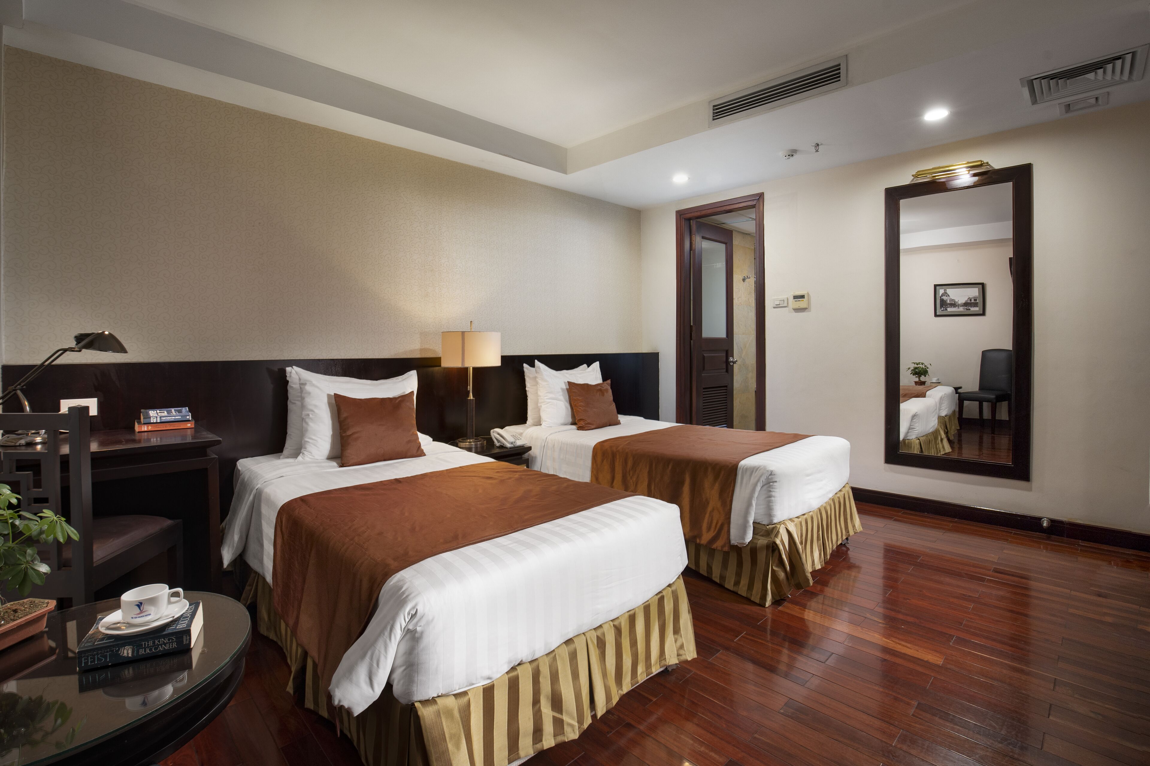 A hotel room with two beds, a round table with a cup and books, a mirror, and a door leading to another room.