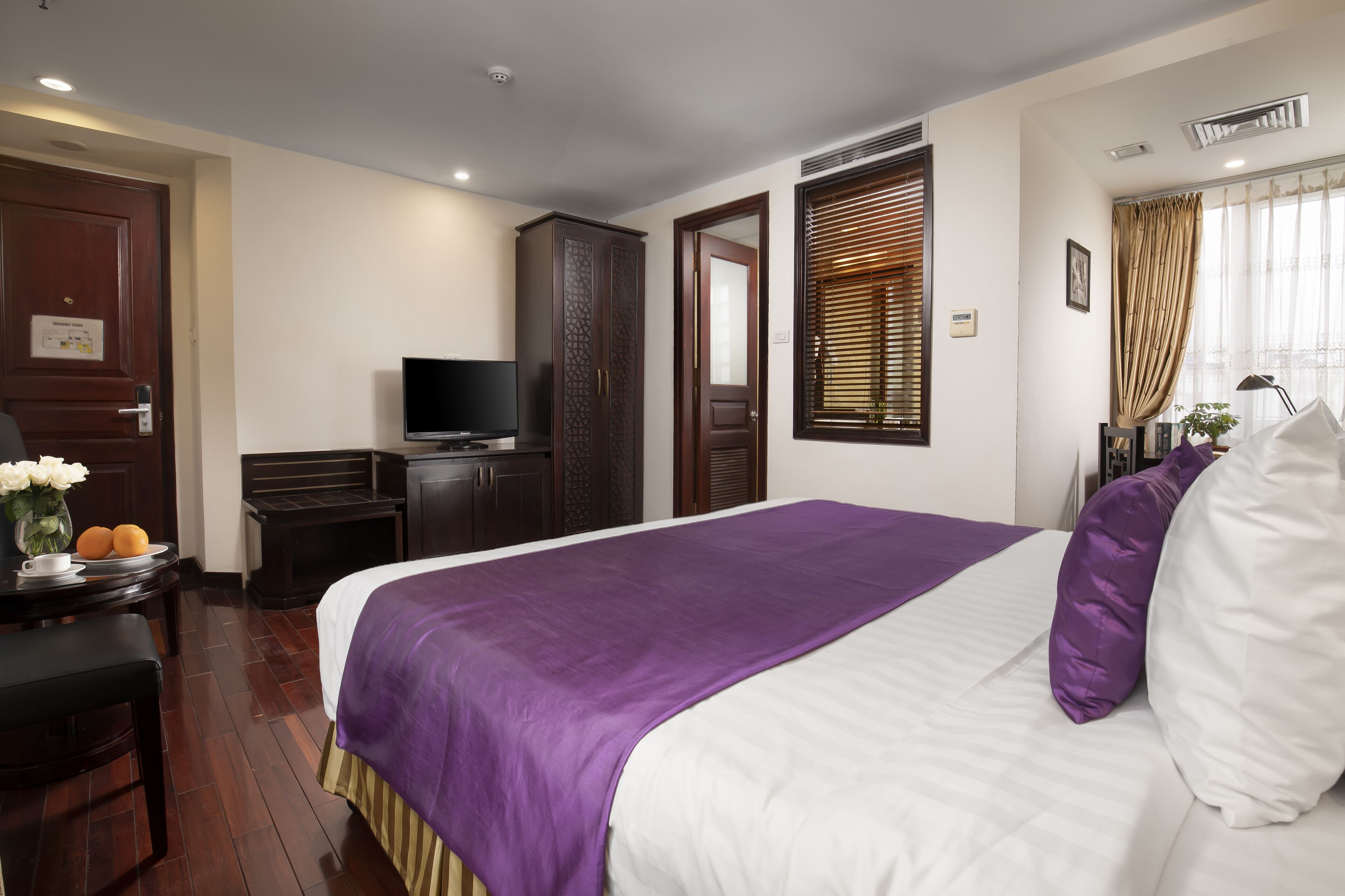 A hotel room with a large bed, a television, a wardrobe, and a desk with a vase of flowers.