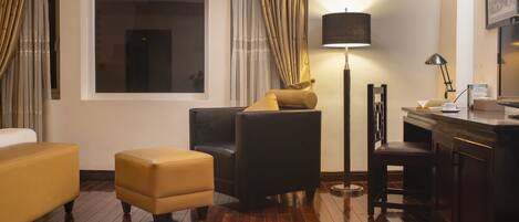 A hotel room with a large window, a sofa, a chair, a desk, and a lamp.