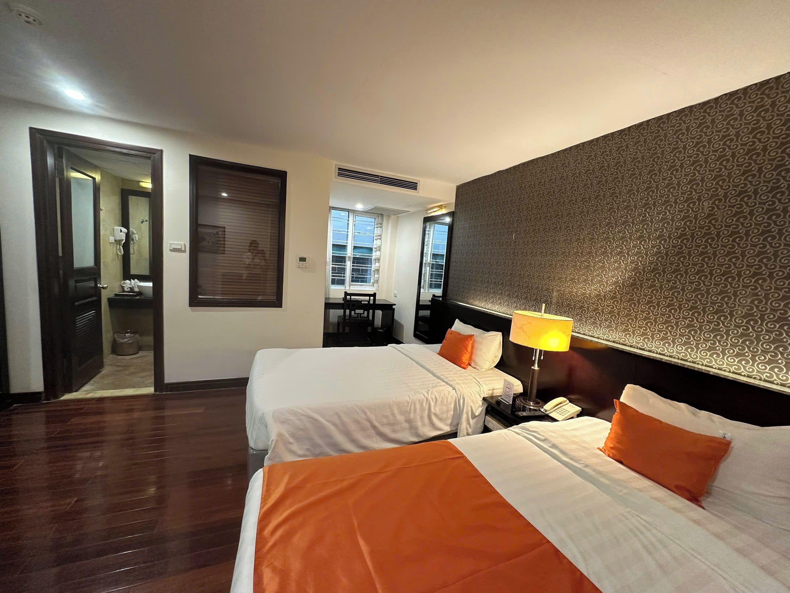 A hotel room with two beds, a desk, a chair, a lamp, and a bathroom visible through an open door.