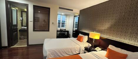 A hotel room with two beds, a desk, a chair, a lamp, and a bathroom visible through an open door.